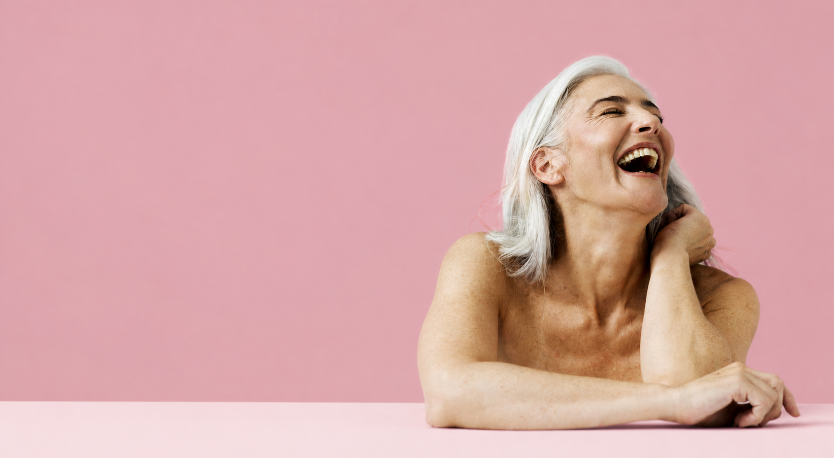 A smiling gray-haired woman laughing against a pink background.