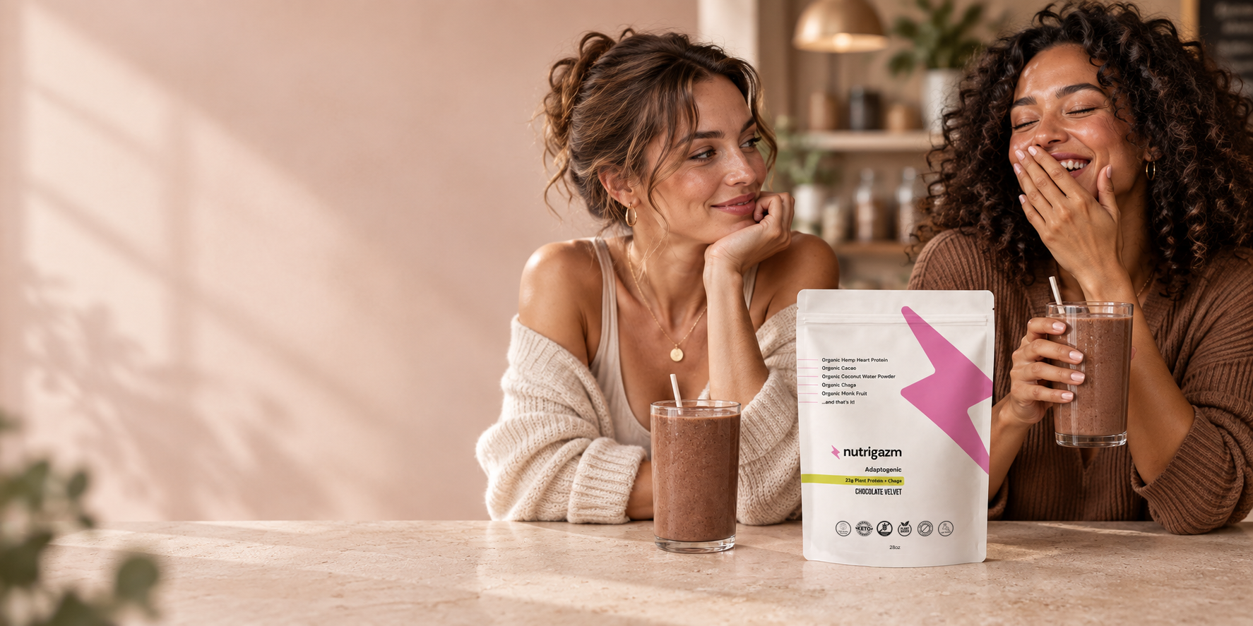 Two women laughing together over Nutrigazm shakes beside a Chocolate Velvet pouch.
